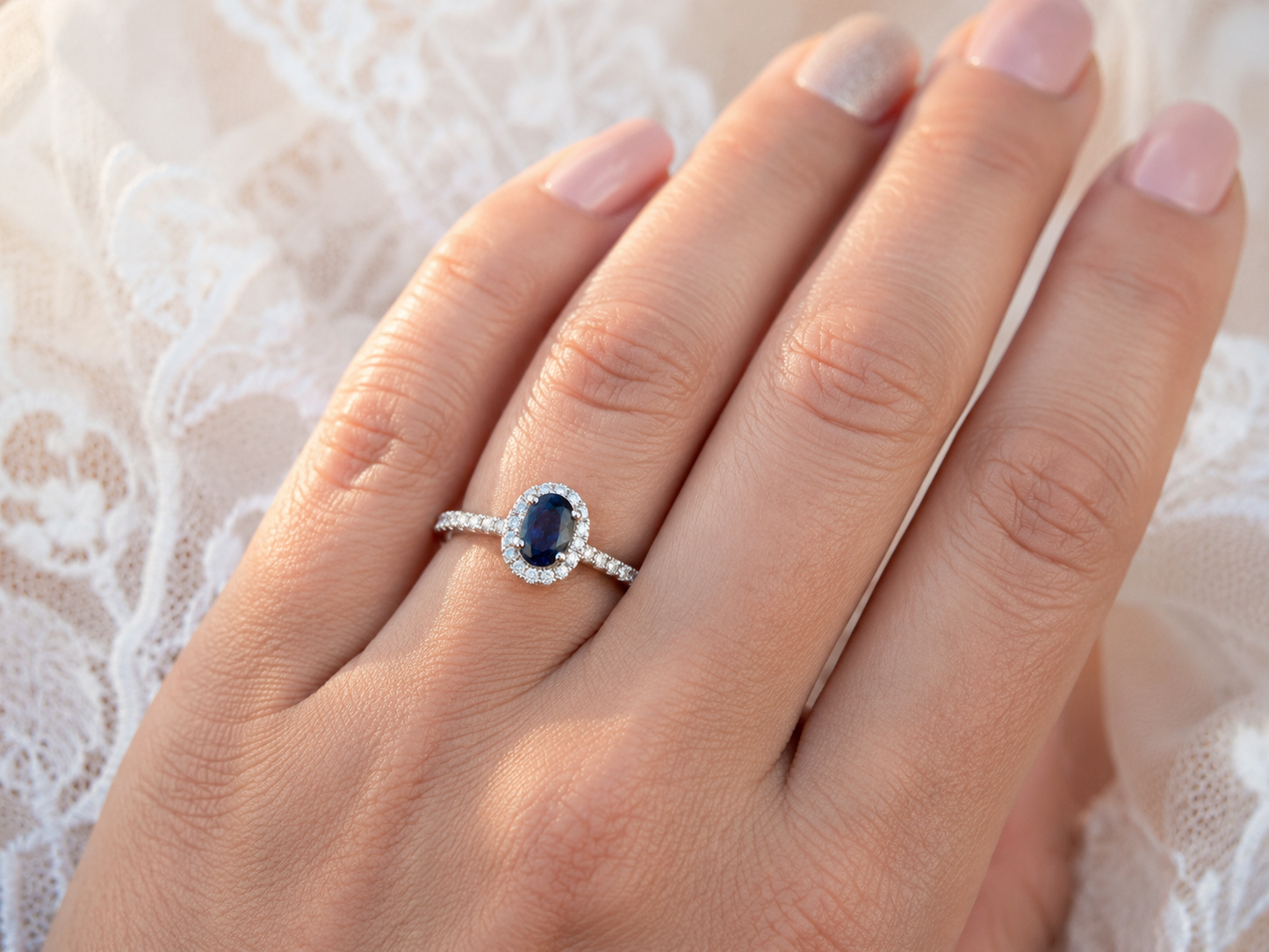 Gros plan sur une main de femme portant une bague de fiançailles en saphir bleu ovale entouré de diamants, façonnée en or blanc 18 carats.