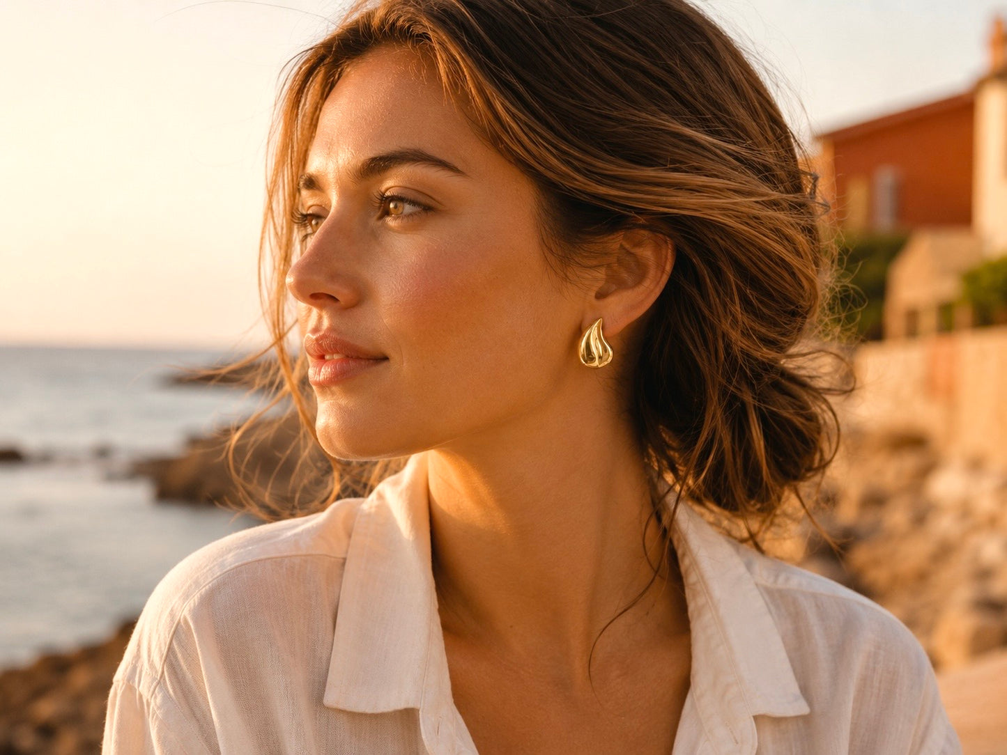 Boucles d'oreilles gouttes d'eau en or jaune 18 carats portées par une femme de profil habillée de blanc au crépuscule.