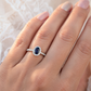 Gros plan sur une main de femme portant une bague de fiançailles en saphir bleu ovale entouré de diamants, façonnée en or blanc 18 carats.