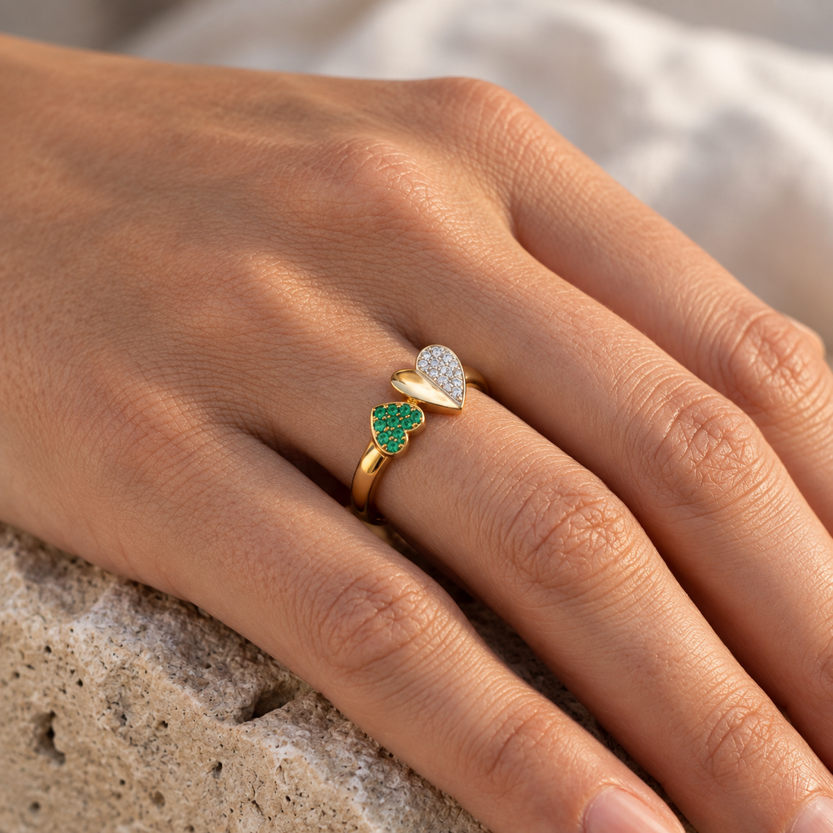Bague Toi et Moi en or jaune 18 carats ornée d'un double coeur en diamants et émeraude verte, portée au doigt d'une femme au soleil.