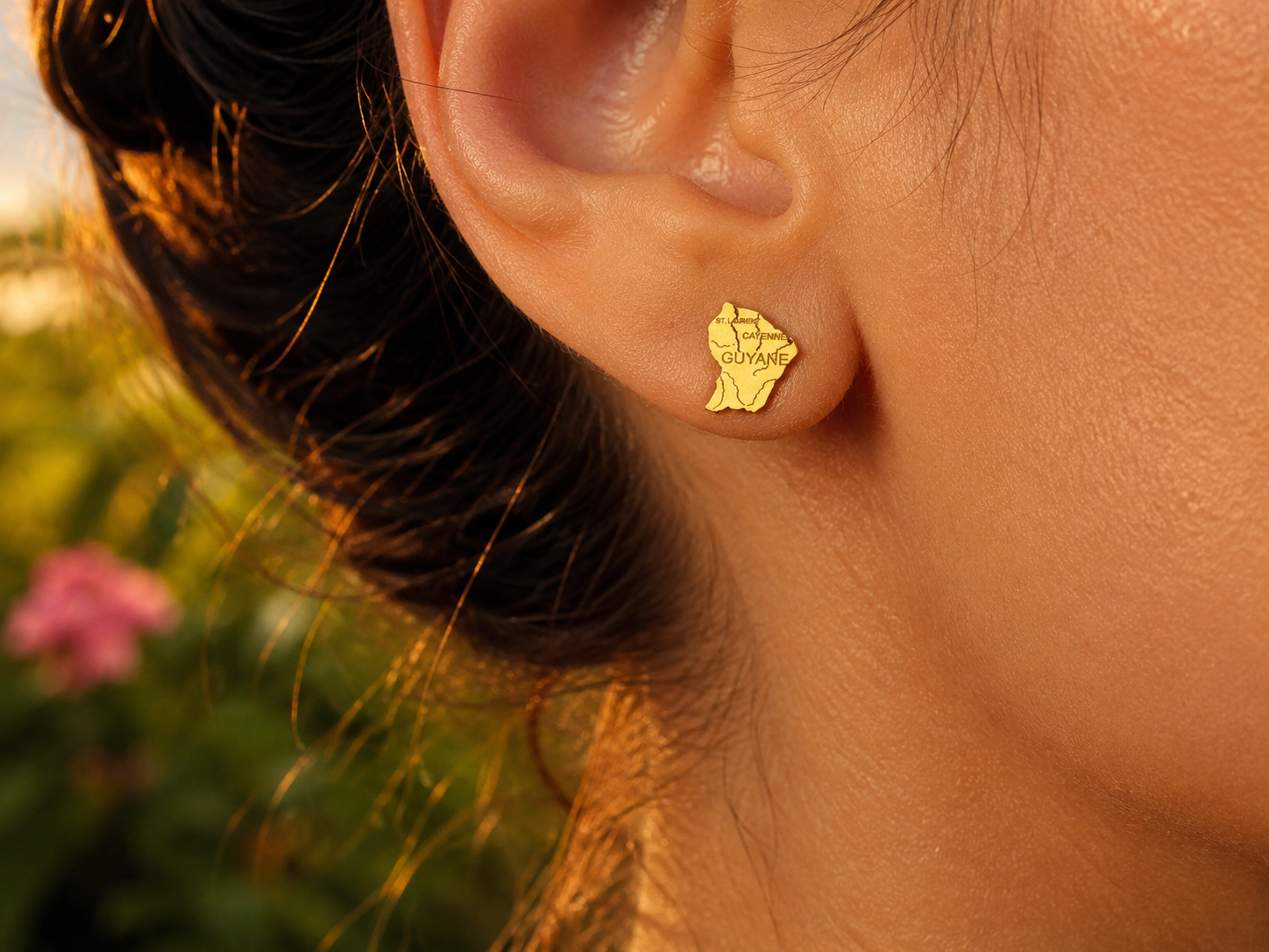 Gros plan sur une oreille de femme portant une puce d'oreille en or jaune 18 carats en forme de carte de la Guyane, bijou de 10x8 mm.