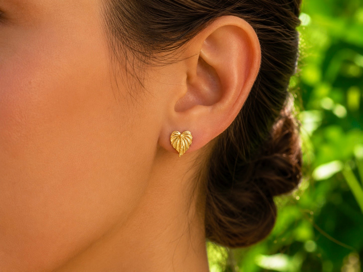 Gros plan sur une oreille de femme portant des boucles d'oreilles en forme de feuille d'Anthurium en or jaune 18 carats, un bijou botanique élégant et lumineux.