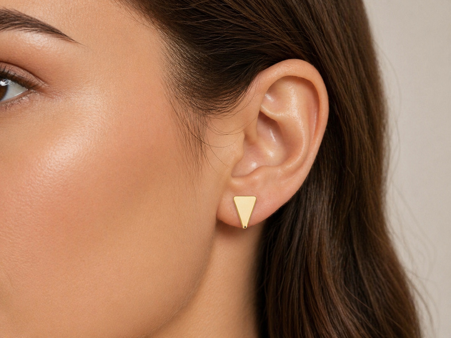  Gros plan sur une oreille de femme portant des puces d'oreilles triangle en or jaune 18 carats, design géométrique et minimaliste.