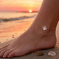 Gros plan sur la cheville d'une femme portant une chaîne en or 18 carats avec un trèfle en nacre, marchant sur le sable au coucher du soleil.