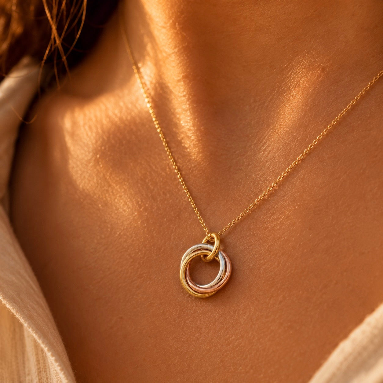 Pendentif trois anneaux entrelacés en or jaune, rouge et gris porté sur une chaîne en or 18 carats au soleil.