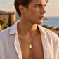 Collier avec pendentif lingot d'or 18 carats porté par un homme en chemise de lin au crépuscule.
