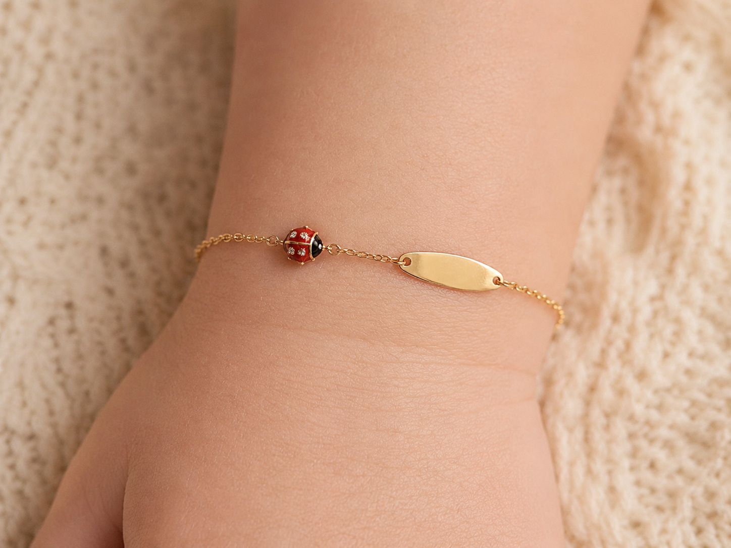 Gros plan sur le poignet d'un bébé portant une gourmette coccinelle rouge en or jaune 18 carats avec plaque ovale lisse.