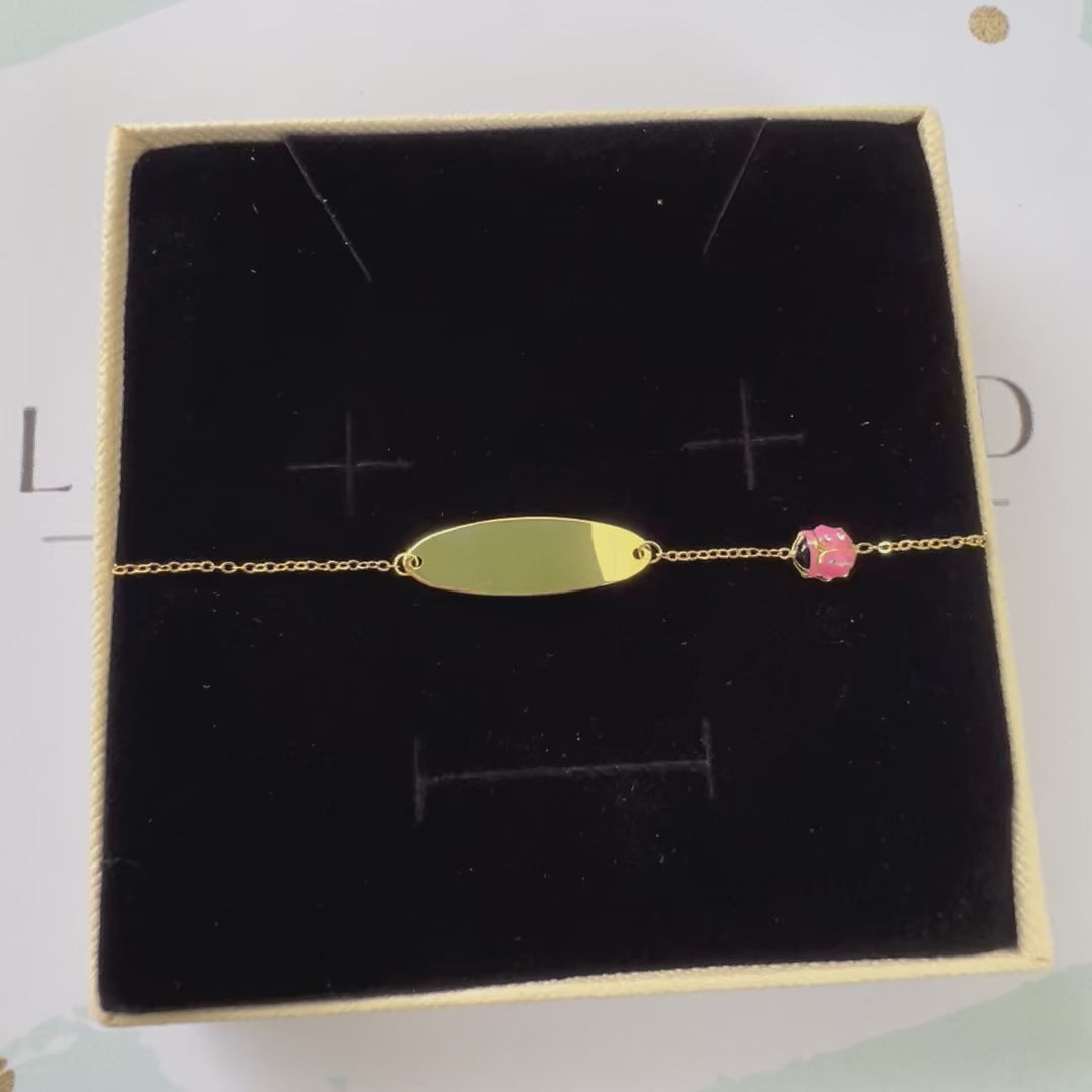 Vidéo de la gourmette de baptême pour bébé en or jaune 18 carats, ornée d'une plaque à graver et d'une petite coccinelle en résine rose sertie d'oxydes de zirconium, délicatement posée dans son écrin noir.