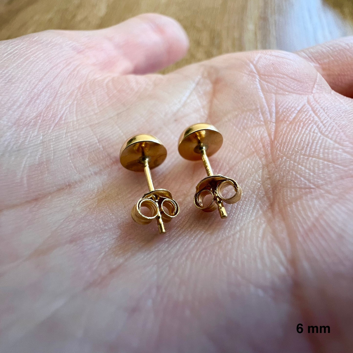 Aperçu de la taille réelle des puces d'oreilles demi-boule en or 18 carats de 6 mm dans la paume d'une main avec leurs fermoirs belge